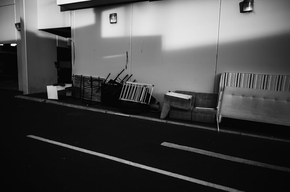 An outdoor scene featuring a beige upholstered sofa with a quilted pattern on the backrest, positioned against a plain white wall. The sofa appears to be in a weathered condition, with some visible st