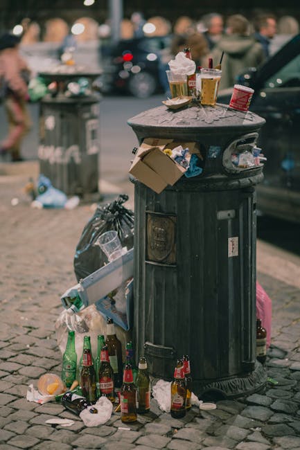 A middle-aged man wearing a black T-shirt with white text is seen disposing of a white plastic garbage bag into a metallic recycling bin situated on a paved walkway. His left hand holds the top of the